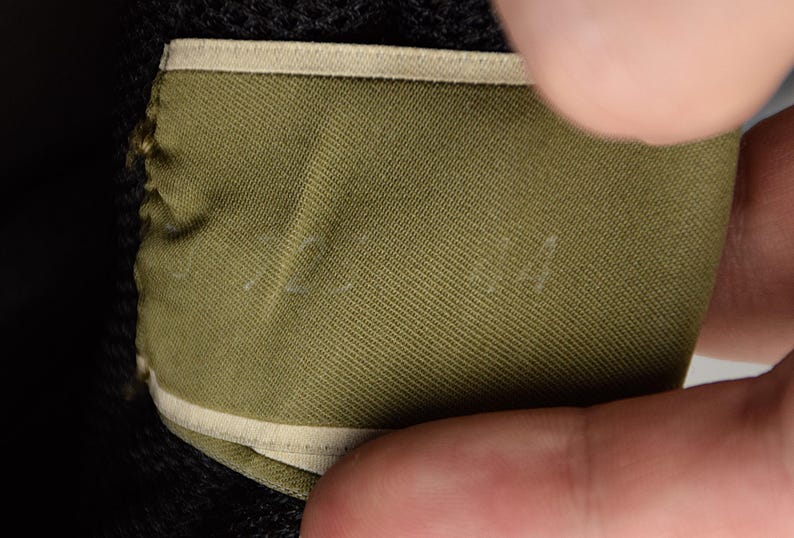 Puede incluir: Primer plano de una etiqueta de tela verde con un borde beige. La etiqueta est&aacute; parcialmente oscurecida por una mano. La tela tiene una apariencia texturizada. Texto blanco tenue visible en la etiqueta.