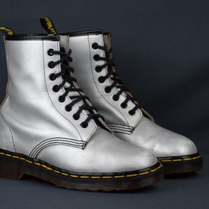 Puede incluir: Un par de botas plateadas con cordones negros y costuras amarillas alrededor de las suelas. Las botas tienen un diseño clásico, con una suela marrón oscuro.