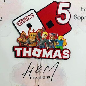 May include: A custom cake topper featuring the Roblox logo, the number 5, and the name THOMAS in bold white letters with red outlines. The design includes five colorful character figures and is attached to a clear plastic stick for placement.