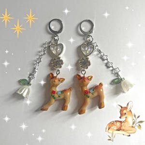 May include: A pair of dangle earrings featuring small deer figurines with floral accents. The earrings have silver-tone hoops, heart-shaped charms, and star and flower details. Each earring has a white flower charm at the end.