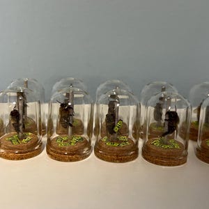 May include: A collection of miniature glass bell jars, each containing a small, detailed figure on a cork base. The figures appear to be insects or small creatures, with green and yellow accents. The jars are arranged in a row.