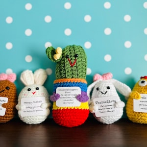 May include: Five crocheted plushies with embroidered faces and motivational messages. The plushies are a potato, two rabbits, a cactus, and a chick. The potato is brown, the rabbits are white, the cactus is green, and the chick is yellow.