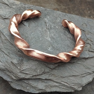 May include: A copper-toned cuff bracelet with a twisted design. The metal is formed into a continuous spiral. The bracelet has a polished finish, reflecting light. The ends are open, allowing it to be slipped over the wrist.