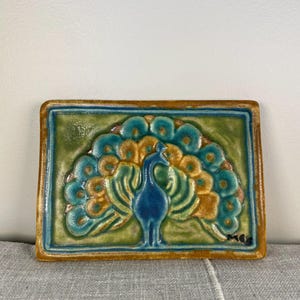 Puede incluir: Azulejo cerámico rectangular con diseño de pavo real. El pavo real tiene cuerpo azul y cola en abanico en tonos turquesa y dorado. El azulejo tiene fondo verde y borde marrón. Mide unos 15 cm de largo.