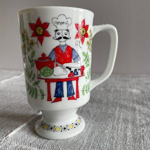 May include: White ceramic mug with a chef illustration. The chef wears a red vest, blue pants, and a chef's hat with the word "mons". The mug features red flowers and green foliage. The mug has a pedestal base and a white handle.