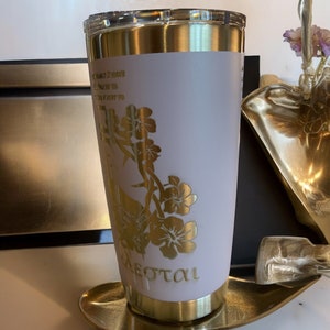 May include: A gold and blush pink stainless steel tumbler with a floral design and the word "Sotal" engraved on the bottom.
