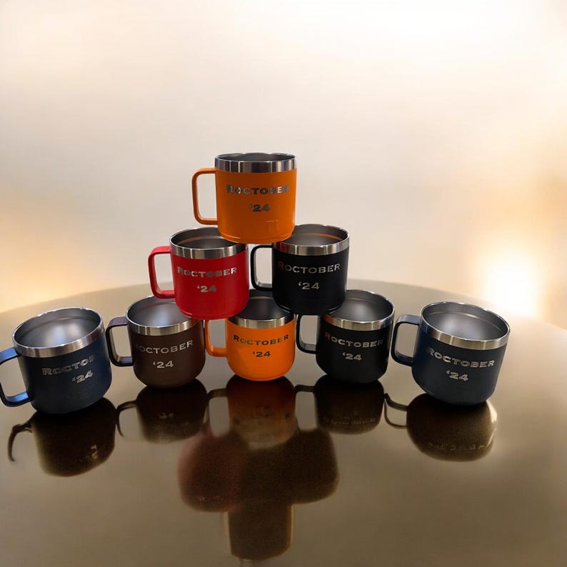 Puede incluir: Ocho tazas de viaje de acero inoxidable en una variedad de colores, incluyendo naranja, rojo, marr&oacute;n, negro y gris. Cada taza tiene el texto "OCTOBER '24" impreso en el lateral.