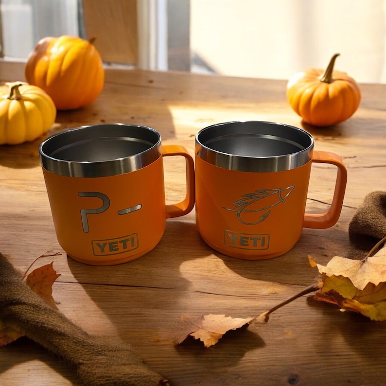 Puede incluir: Dos tazas Yeti Rambler de color naranja con tapas y asas de acero inoxidable. La taza de la izquierda tiene el texto "P- YETI" grabado en el lateral. La taza de la derecha tiene un se&ntilde;uelo de pesca con mosca grabado en el lateral con el texto "YETI".