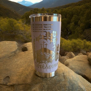 May include: A stainless steel tumbler with a white, lavender-colored design. The tumbler features a gold-colored image of two crosses, a crown of thorns, and the word "Tetelestai" in Greek. The tumbler is sitting on a rock with a mountain range in the background.