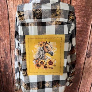 May include: A long-sleeved, button-down flannel shirt with a black and white plaid pattern. The back features a yellow square with a horse head surrounded by sunflowers. The shirt has brown and tan accents.