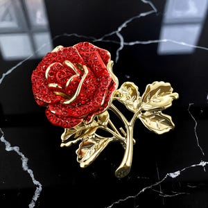 May include: A detailed brooch in the shape of a rose. The rose petals are covered in red crystals, with gold-colored accents and leaves. The brooch is set against a black marble-like surface.