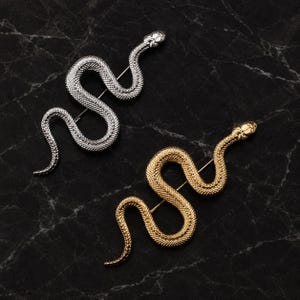 Puede incluir: Dos broches con forma de serpiente, uno en tono plateado pulido y otro en tono dorado. Cada broche presenta un detalle de superficie texturizada similar a escamas a lo largo del cuerpo y una cabeza definida, con un mecanismo de alfiler visible.