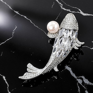 May include: A silver-toned fish brooch with a pearl and clear crystal accents. The fish has a pearl near its head and is covered in small, sparkling crystals. The body of the fish is made of larger, clear crystals. The brooch is set against a black background.