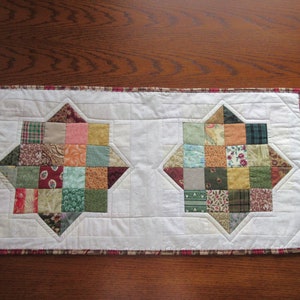 Peut inclure: Un chemin de table blanc avec une bordure rouge présente deux motifs d'étoiles en patchwork. Les étoiles sont faites de différents tissus dans des tons de vert, marron, orange et rose.