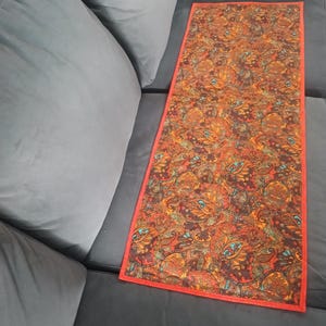 May include: A rectangular table runner with a red border. The runner has a paisley pattern in brown, orange, and teal. It is placed on a dark grey sofa. The runner's length is approximately 90 cm.