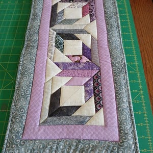 May include: A handmade quilted table runner with a geometric design. The runner features a central pattern of squares and triangles in shades of purple, grey, and white, framed by a lavender border and a grey patterned edge.