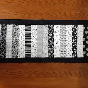 Peut inclure: Chemin de table rectangulaire avec une bordure noire et un motif central de bandes de tissu verticales. Les bandes présentent divers motifs noirs et blancs, notamment des motifs floraux, cachemire et géométriques. Le chemin de table est posé sur une surface en bois.
