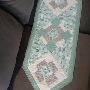 May include: A rectangular table runner with a light teal background featuring a repeating pattern of cat faces and the word "MEOW". The runner has three square patchwork designs in white, grey, and teal. The edges are trimmed with a teal border.