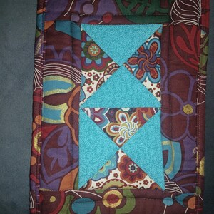 May include: A quilted fabric item with a brown background and a central design of turquoise triangles. The background features a floral pattern in shades of red, blue, and green. The edges are neatly stitched.
