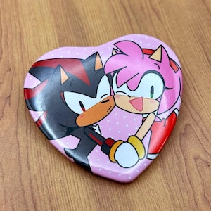May include: Heart-shaped pin featuring the characters Shadow and Amy Rose from the Sonic the Hedgehog series. The pin has a pink background with white polka dots. The characters are hugging and kissing.