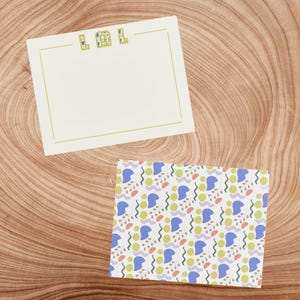 Può includere: Un biglietto bianco vuoto con la parola "LOL" stampata in lettere verdi in alto. Il biglietto è su una superficie di legno. Una busta a motivi con sfondo bianco e forme blu, verdi, gialle e arancioni è anche sulla superficie.