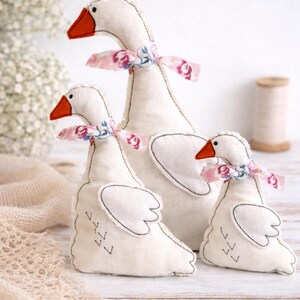 May include: Three white goose plush toys with orange beaks and floral patterned ribbon bows around their necks. The geese have black stitched details and are arranged in a group, varying in size. The background is a soft, neutral color.