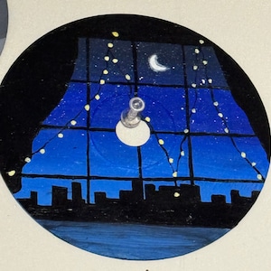 May include: A painted vinyl record with a night scene of a city skyline viewed through a window with string lights. The sky is blue with a crescent moon and stars.