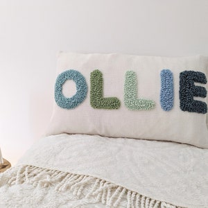 May include: A rectangular cushion embroidered with the name "OLLIE" in textured letters. The letters are in shades of blue and green, set against a cream background. The cushion rests on a bed with a white blanket.