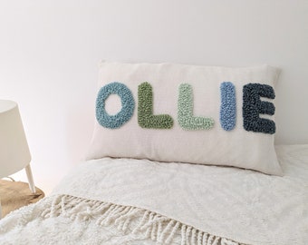May include: A rectangular pillow with the name "OLLIE" embroidered in textured, colorful letters. The letters are in shades of blue and green, set against a cream-colored background. The pillow is placed on a bed with a white blanket.