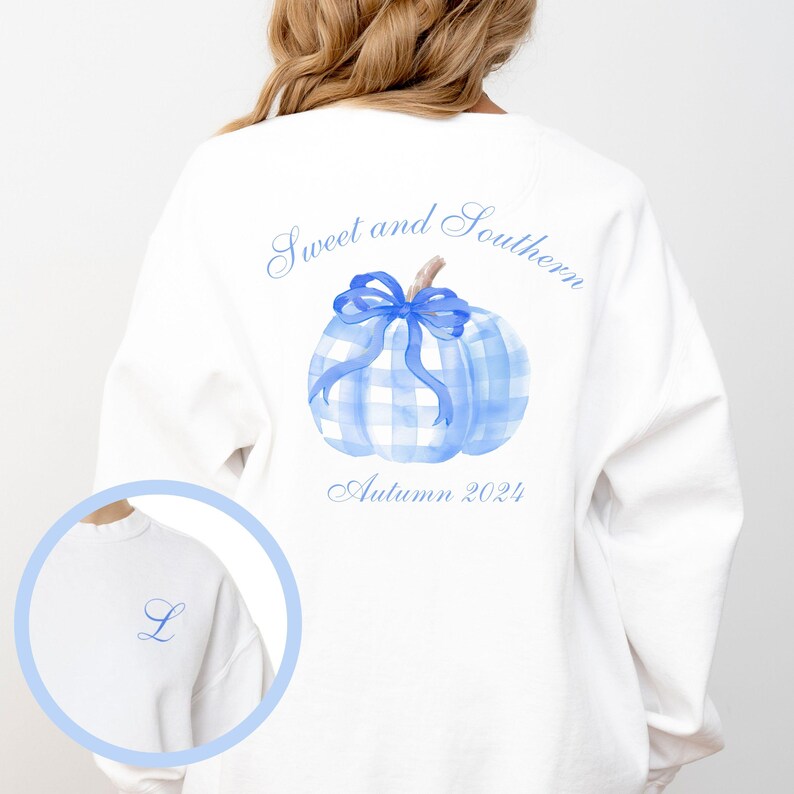 Op de afbeelding: Witte sweatshirt met een blauw geruit pompoenontwerp, een strik en de tekst "Sweet and Southern" en "Autumn 2024". Een kleine blauwe "L" staat op de voorkant. Het ontwerp is in aquarel stijl.