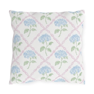 Puede incluir: Cojín decorativo cuadrado con fondo blanco que presenta un patrón repetido de flores de hortensia azules y hojas verdes. Las flores están dispuestas dentro de un diseño de celosía rosa.