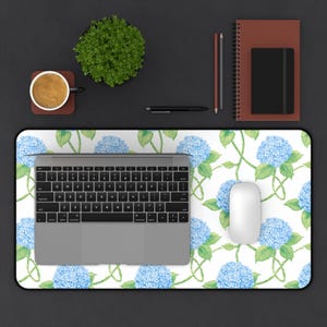 May include: A floral desk mat featuring a pattern of blue hydrangea flowers and green vines on a white background. A laptop, mouse, coffee cup, and other office supplies are arranged on the mat.
