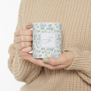 Op de afbeelding: Witte keramische mok met een blauw en groen bloemenpatroon en een wit vierkant met de letter "F" in een cursief schrift.