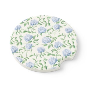 May include: Round car coaster with a floral design. The coaster features a cream-colored background with a pattern of blue hydrangea flowers and green vines. The coaster has a small notch on one side for easy removal from a car cup holder.