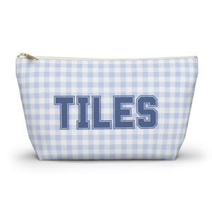 May include: A light blue and white gingham patterned zippered pouch with the word "TILES" in navy blue block letters. The pouch has a gold-colored zipper and is likely used for cosmetics or small items.