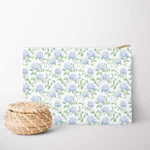 Puede incluir: Un estuche rectangular con cremallera con un patrón repetido de flores de hortensia azules y hojas verdes sobre un fondo blanco. El estuche tiene una cremallera dorada. Hay una pequeña cesta tejida al lado.