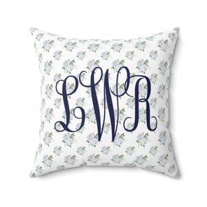 以下が含まれることがあります： 白い背景に小さな青い花のブーケの模様が入った正方形のクッション。クッションには、L、W、Rの文字が装飾的な筆記体で大きくネイビーブルーのモノグラムが施されています。クッションは正方形です。