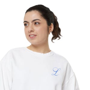 Op de afbeelding: Een witte sweatshirt met een blauwe, cursief geborduurde letter "L" op de voorkant. De sweatshirt heeft een ronde hals en lijkt gemaakt te zijn van een zacht materiaal. De afbeelding is een close-up van de sweatshirt.