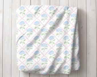 Puede incluir: Tela blanca con un patrón repetido de flores de hortensia azules y enredaderas rosas. La tela está doblada y drapeada sobre una superficie de madera blanca.