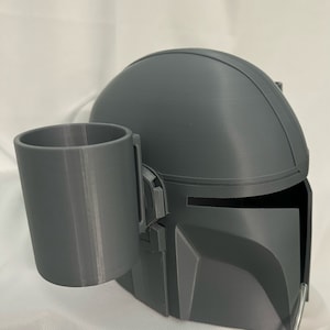 以下が含まれることがあります： グレーの3Dプリントされたマンダロリアンヘルメットとカップホルダー。ヘルメットはバイザーと丸みを帯びたデザインが特徴です。カップホルダーは円筒形で、ヘルメットの左側に配置されています。