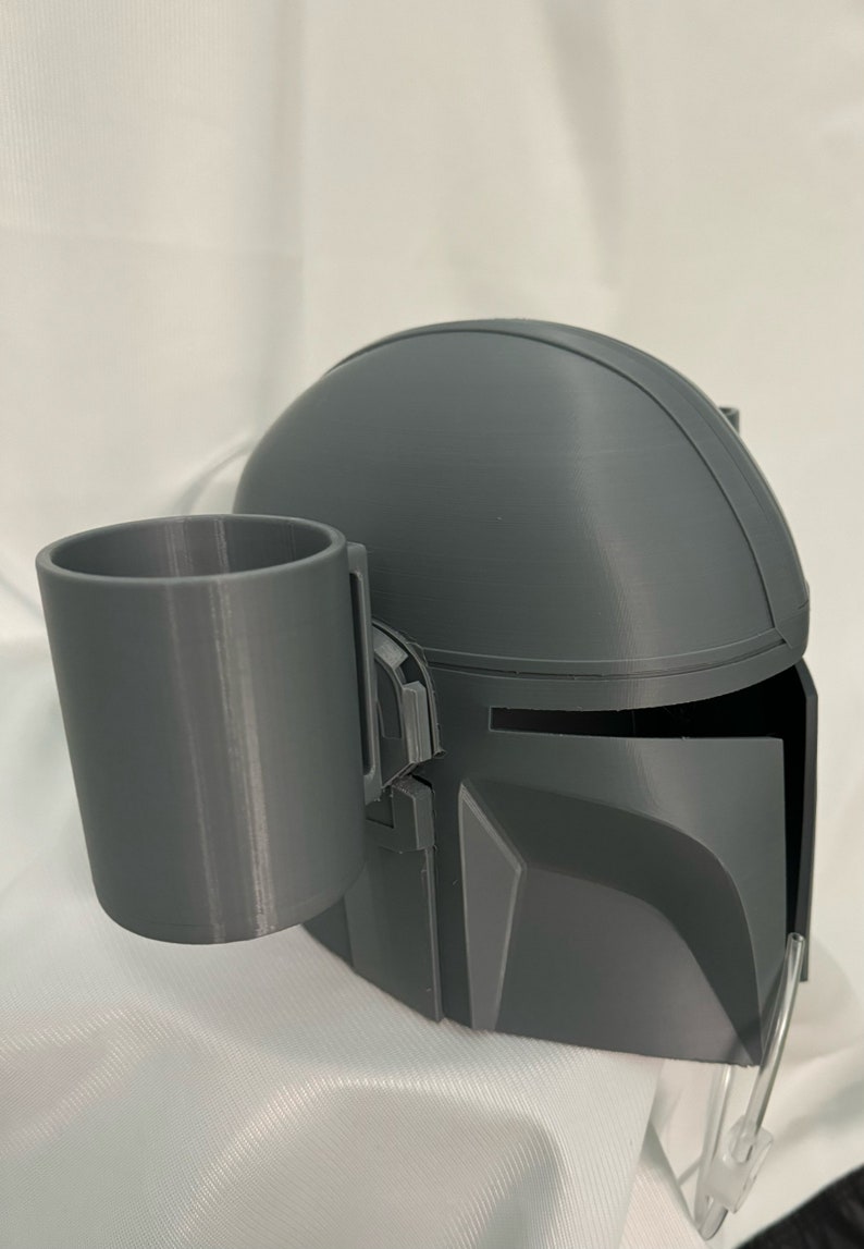 以下が含まれることがあります： グレーの3Dプリントされたマンダロリアンヘルメットとカップホルダー。ヘルメットはバイザーと丸みを帯びたデザインが特徴です。カップホルダーは円筒形で、ヘルメットの左側に配置されています。