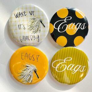 May include: Four round buttons with text and graphics. One button has the text "WAKE UP IT'S GAMEDAY" with a yellow and white checkered background. Another button has the word "Eags" in white on a black background with yellow polka dots. The other two buttons have the word "Eags" on a yellow background.