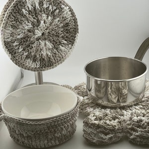 Puede incluir: Tres posavasos de ganchillo en color beige y blanco. Un posavasos es redondo y está en un soporte, otro es redondo y está en un cuenco de cerámica blanco, y el tercero tiene forma de estrella y está debajo de una olla de plata.