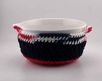 May include: A white ceramic bowl with a navy blue, red, and white crocheted cozy. The cozy has a striped pattern and handles. The bowl is likely for serving or holding food.