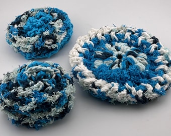 May include: Three handmade crochet scrubbies in shades of blue, white, and gray. The scrubbies are round and have a textured appearance, perfect for kitchen or bath use. The largest scrubby is approximately 4 inches in diameter.