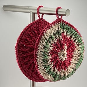 May include: Two handmade, round crochet potholders hanging from a metal bar. One is solid burgundy, the other features a spiral design in burgundy, cream, and green. The potholders are made of thick yarn and have a textured appearance.