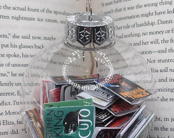 May include: A clear glass Christmas bauble filled with miniature book covers, including Stephen King's "Cujo" and "The Shining." The bauble has a silver cap and a silver string for hanging. The background is an open book.