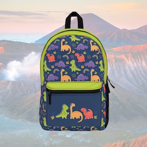 Puede incluir: Una mochila azul marino con ribete verde y asa negra. La mochila presenta un colorido estampado de dinosaurios con dinosaurios naranjas, verdes, morados y rojos. Un bolsillo más pequeño en la parte delantera tiene una fila de dinosaurios.