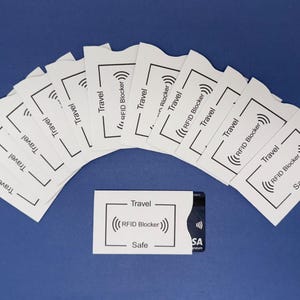 Op de afbeelding: Een set van tien witte RFID-blokkeerhoesjes voor kaarten met de tekst "Travel" en "RFID Blocker" erop gedrukt. De hoesjes zijn ontworpen om creditcards en andere gevoelige kaarten te beschermen tegen elektronische diefstal.