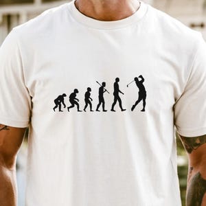 May include: A cream-colored t-shirt featuring a black graphic of the evolution of a golfer. The design shows a progression from ape-like figures to a golfer in mid-swing. The text "Tshirts & Sweatshirts" is at the bottom.